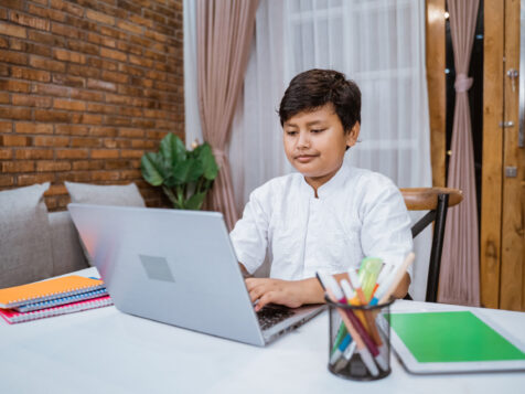 Cheerful boy writing using laptop while doing schoolwork memorize-quran-online-in-italy